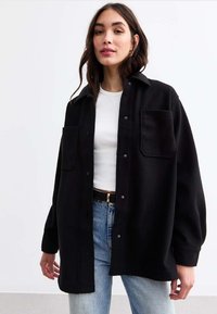 Giacca camicia nera oversize con due tasche frontali, chiusura a bottoni, realizzata in tessuto spesso, indossata sopra una maglietta bianca e abbinata a jeans azzurri chiari.