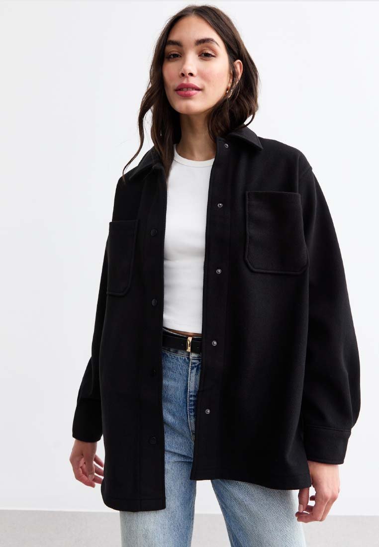 Giacca camicia nera oversize con due tasche frontali, chiusura a bottoni, realizzata in tessuto spesso, indossata sopra una maglietta bianca e abbinata a jeans azzurri chiari.