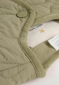 Gepunktete, olivgrüne Babyjacke mit rundem Kragen, Druckknopfverschluss und weicher Innenauskleidung. Das Etikett zeigt ein Sternenmotiv.