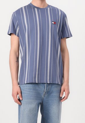 Homme portant un t-shirt bleu à manches courtes avec des rayures verticales blanches et un petit logo Tommy Jeans sur la poitrine, associé à un jean bleu clair.