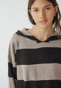 Haut à capuche rayé noir et beige avec une texture douce, présentant une coupe décontractée et une capuche avec cordon de serrage. Les accents incluent de larges rayures et une encolure arrondie.