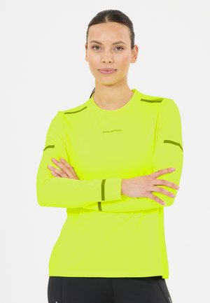 Vrouw met donker haar in een staart draagt een felgeel lange mouwen sportshirt met zwarte strepen en zwarte broek, staand met gekruiste armen.