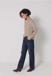 Jersey beige de punto con cuello alto, combinado con jeans rectos de color azul oscuro y botas de tobillo marrones, de pie contra un fondo simple.