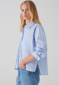 Camicia a righe azzurro chiaro in cotone con maniche lunghe, colletto con bottoni, taschino singolo sul petto e polsini arrotolati.