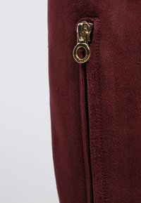 Bolso de ante burdeos con un zipper dorado. Superficie texturizada con detalles de costura a lo largo del borde, con un acento de tirador de zipper redondo.