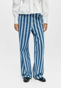 Personne portant un jean taille haute à jambes larges avec des rayures bleues verticales, associé à un chemisier blanc à manches longues et des chaussures noires.