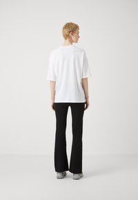 Tricou oversized din bumbac alb, cu mâneci scurte, asociat cu pantaloni negri evazați. Modelul stă cu spatele la cameră, pe un fundal neutru.