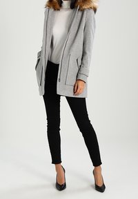 Grå frakke i uldblanding med pelsforet hætte, sidelommer og lynlåslukning, kombineret med sort tætsiddende bukser og sorte høje hæle.