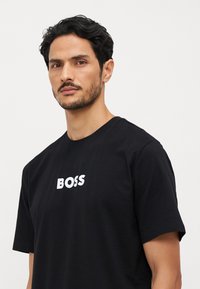 Musta puuvillane T-särk, millel on ümmargune kaelus ja lühikesed varrukad, esitades esiosas selgelt sõna "BOSS" valge värviga.