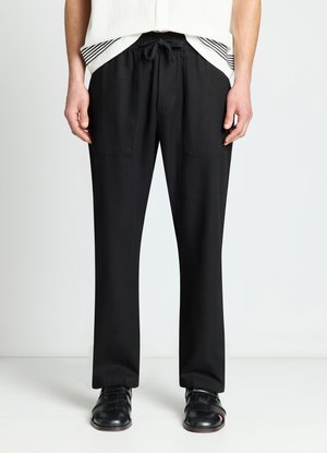 Uomo che indossa pantaloni neri con coulisse e tasche applicate frontali e sandali di pelle nera intrecciata, in piedi su sfondo bianco.