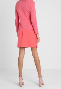 Kobieta stoi tyłem, ubrana w różową dopasowaną kurtkę, koralową spódnicę z bocznym rozcięciem oraz różowe sandały na obcasie z paskiem wokół kostki.