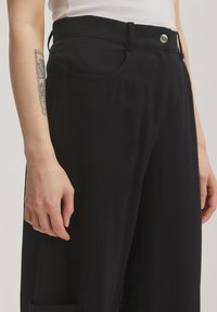Schwarze, hochtaillierte Hose aus weichem Stoff, mit einem silbernen Knopf, Fronttaschen und geradem Bein. Glatte Textur.