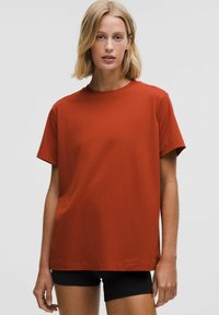 Rūsas krāsas kokvilnas T-krekls ar brīvu piegriezumu, apaļu kakla izgriezumu, īsām piedurknēm un gludu tekstūru. Valkāts ar melnajām šortiem.