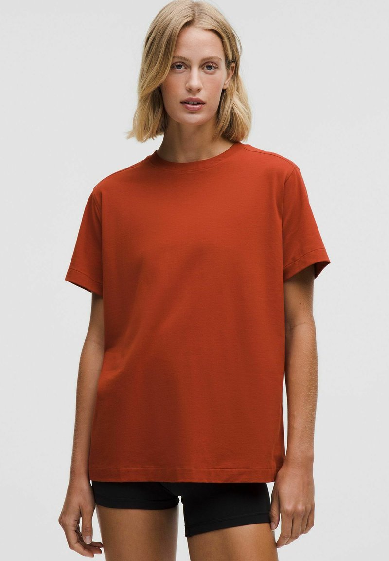 Rūsas krāsas kokvilnas T-krekls ar brīvu piegriezumu, apaļu kakla izgriezumu, īsām piedurknēm un gludu tekstūru. Valkāts ar melnajām šortiem.