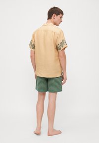 Camisa de lino beige de manga corta con detalles bordados en verde en las mangas, combinada con pantalones cortos de cuadros verdes. Descalzo, visto desde atrás.