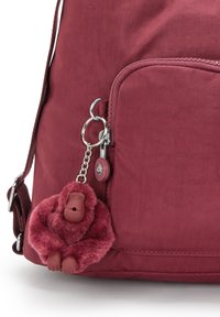 Burgundy ryggsäck med en texturerad tyg, med en sidoficka och en plyschapa nyckelring fäst med en silverkedja.