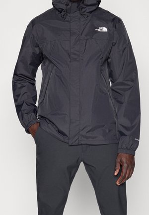 Homme portant une veste de pluie noire à capuche avec des poignets élastiques, des poches zippées et le logo The North Face sur la poitrine, associée à un pantalon noir.