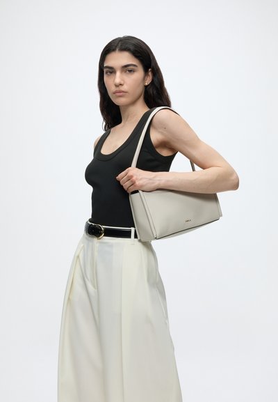 Femme aux longs cheveux foncés tenant un sac à main beige clair sur l'épaule, portant un haut noir sans manches, un pantalon blanc taille haute et une ceinture noire sur fond blanc.