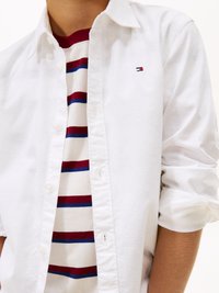 Biała koszula z guzikami założona na kremowy sweter w paski z czerwonymi i niebieskimi liniami. Koszula ma małe logo na piersi.