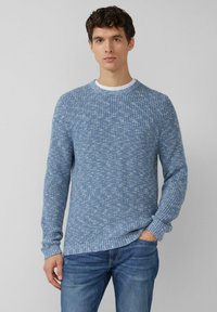 Blauer gestrickter Pullover mit strukturiertem Muster, rundem Ausschnitt und gerippten Bündchen. Trägt sich über einem weißen T-Shirt und wird mit blauer Jeans kombiniert.