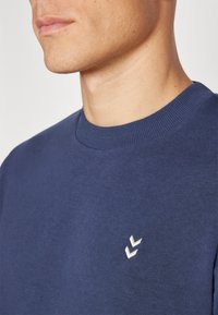 Marineblauwe trui met een ronde hals en geribbelde kraag, voorzien van een klein wit logo op de borst. Zacht materiaal met een gladde textuur.
