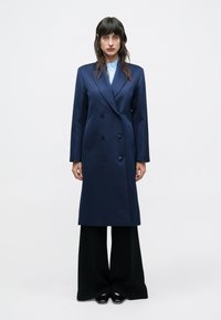 Žena stojaca v tmavomodrom dvojradovom dlhom kabáte, svetlomodrej košeli, čiernych širokých nohaviciach a čiernych špicatých topánkach na jednoduchom pozadí.