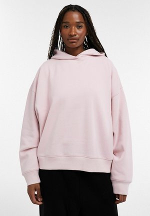 Femme avec de longues tresses portant un sweat à capuche ample rose pâle et un pantalon noir, debout devant un fond blanc uni.