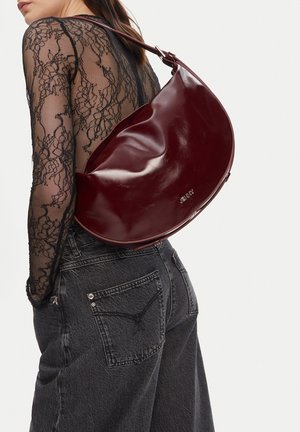 Frau trägt ein schwarzes, transparentes Spitzenoberteil und dunkle Jeans, mit einer großen, glänzenden, dunkelroten Leder-Schultertasche auf dem Rücken.