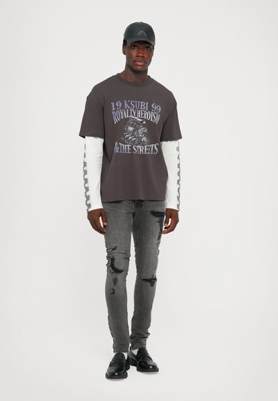 Dunkelgraues T-Shirt mit grafischem Druck, über einem weißen Langarmshirt getragen. Ausgewaschene Skinny-Jeans mit abgetragenen Details. Schwarze Lederschuhe.