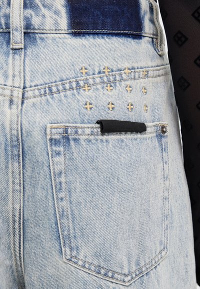 Jeans de mezclilla azul claro con cinturilla azul oscura, con acentos de tachuelas doradas y un detalle de bolsillo negro en la parte trasera. Acabado texturizado.