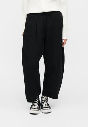 Άτομο που φοράει φαρδιά μαύρη παντελόνα, λευκό μακρυμάνικο μπλουζάκι και μαύρα ψηλά αθλητικά παπούτσια με λευκά κορδόνια, στέκεται σε λευκό φόντο.