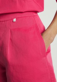 Detailní záběr osoby, která má na sobě jasně růžové kalhoty, ruku v boční kapse a na pásku malou stříbrnou značkovou cedulku.