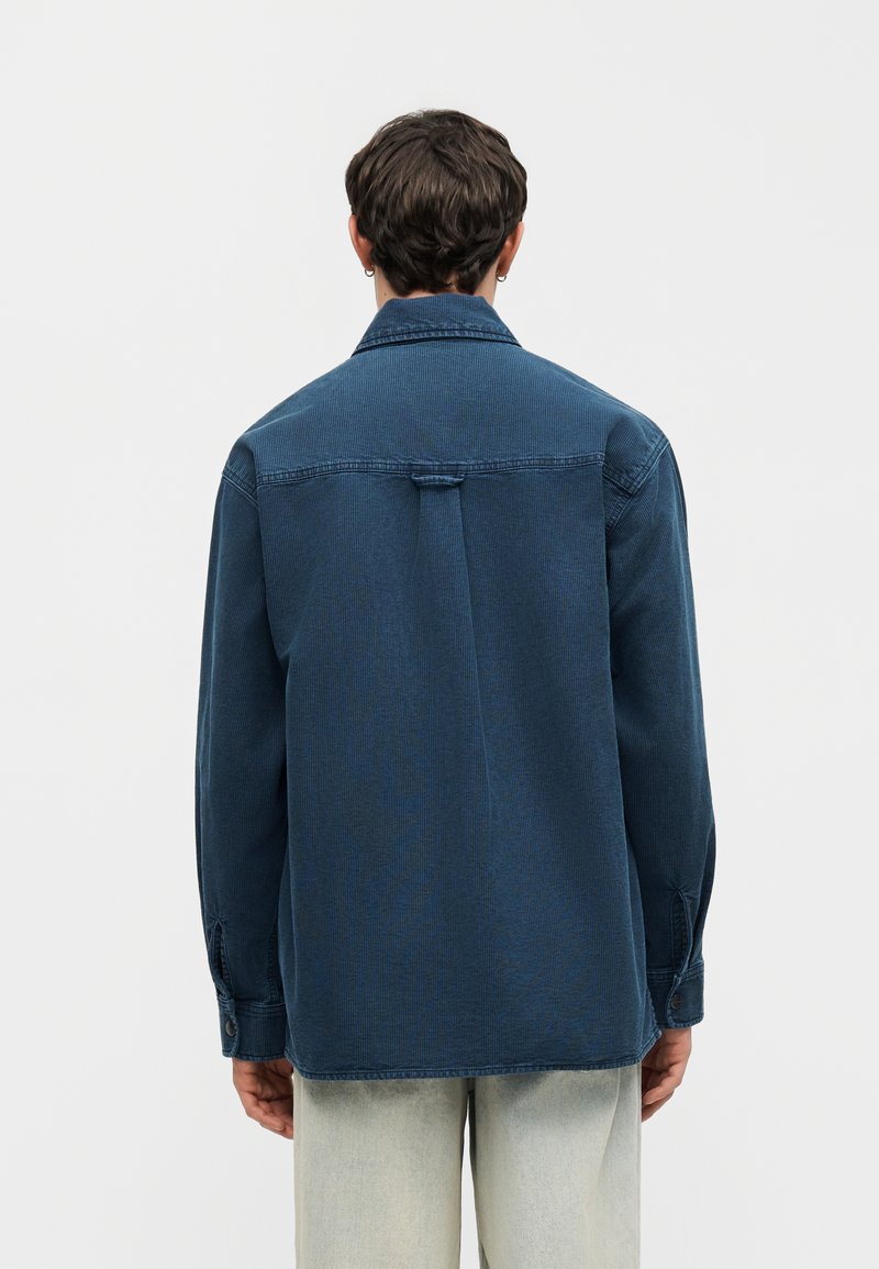 Person mit kurzen Haaren, die ein dunkelblaues, langärmliges Cordhemd und hellfarbene Hosen trägt, von hinten vor einem einfarbigen Hintergrund.