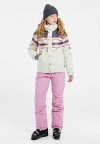 Giacca da sci da donna color crema con motivi geometrici rosa e grigi; abbinata a pantaloni da sci rosa; il modello indossa un berretto a maglia grigio.
