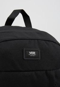Sac à dos noir Vans avec une poignée supérieure et un patch logo cousu portant l'inscription "Vans Off The Wall Since 1966" sur le devant.