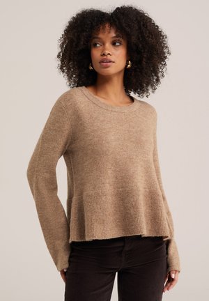 Frau mit lockigem Haar, die goldene Creolen trägt und einen lockeren beigefarbenen Pullover mit einer dunklen Hose kombiniert, steht vor einem schlichten hellen Hintergrund.