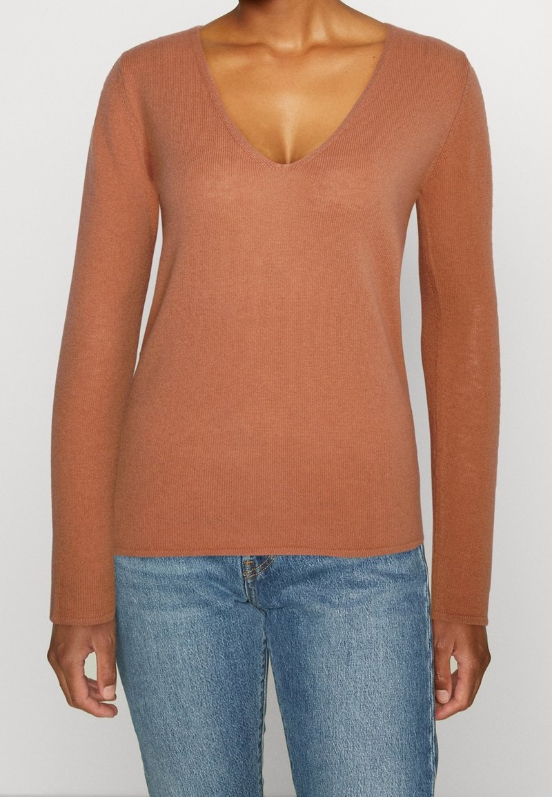 Femme portant un pull cintré à manches longues, de couleur rouille avec un col en V, et un jean bleu, debout devant un fond blanc uni.