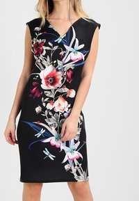 Kvinna i en svart ärmlös klänning med en färgglad blommig och odöda tryck, står framför en enkel ljus bakgrund.