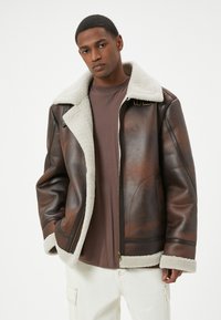 Giacca in pelle marrone con colletto e polsini in shearling, caratterizzata da chiusura con zip e doppi dettagli a fibbia. Finitura testurizzata con dettagli a contrasto.