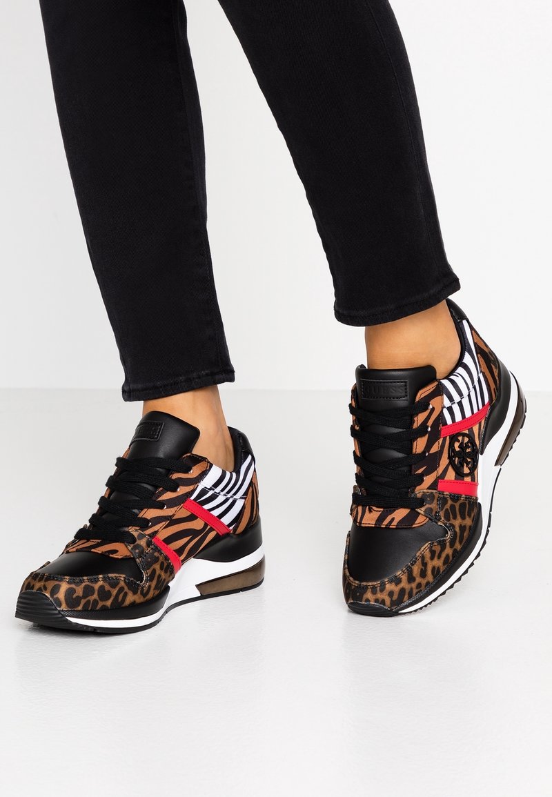 Pieds portant un pantalon noir et des baskets à imprimé animal avec des motifs léopard et zèbre, lacets noirs et accents rouges sur fond blanc.