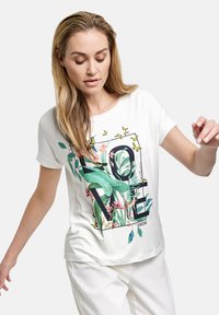 Hvid kortærmet t-shirt med et farverigt botanisk print, der har teksten "KÆRLIGHED", grønne blade og et dovendyr som accent. Blødt, letvægts stof.