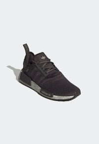 Svarta Adidas NMD-sneakers med ovandel i stickat, reflektiva snören, grå texturerad mellansula och märkt dragflik på vit bakgrund.