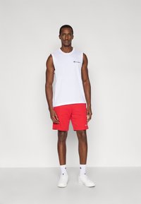 Λευκό αμάνικο αθλητικό μπλουζάκι με μικρό μαύρο λογότυπο, σε συνδυασμό με κόκκινα σορτς. Λευκά αθλητικά παπούτσια και λευκές κάλτσες με λογότυπο. Απλό φόντο.