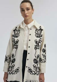 Knoop-up crème cardigan met zwarte bloemenborduursels, textuurstof en een franjeszoom. Gestyled over een witte shirt. Zwarte knopen zijn zichtbaar.