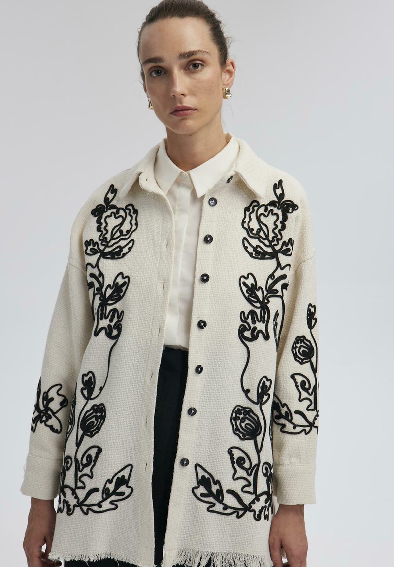 Knoop-up crème cardigan met zwarte bloemenborduursels, textuurstof en een franjeszoom. Gestyled over een witte shirt. Zwarte knopen zijn zichtbaar.
