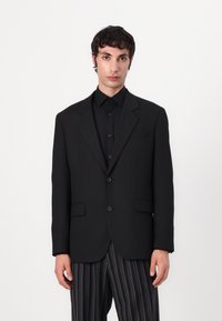 Black tailored blazer with two buttons, notch lapels, and two front pockets, paired with a black shirt and striped pants. Smooth texture.