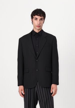 Sacou negru ajustat cu două butoane, rever tip notch și două buzunare frontale, asortat cu o cămașă neagră și pantaloni lungi cu dungi. Textură netedă.