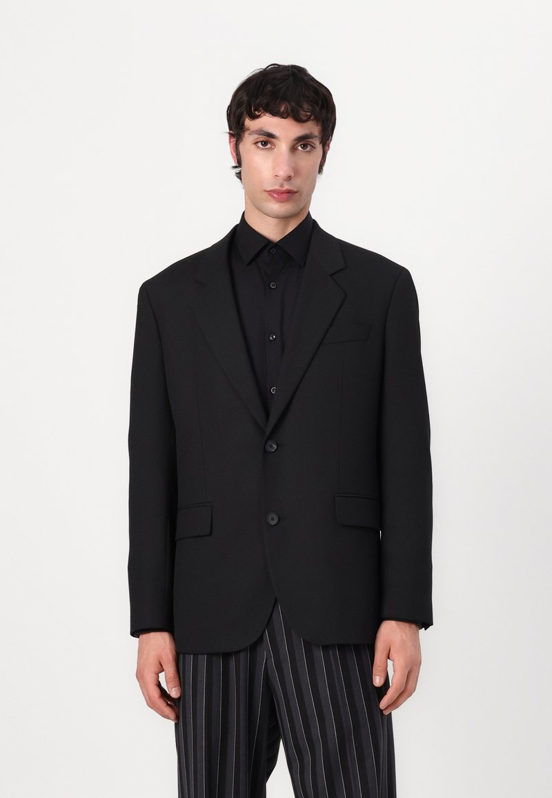 Black tailored blazer with two buttons, notch lapels, and two front pockets, paired with a black shirt and striped pants. Smooth texture.