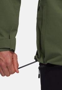 Veste verte avec une texture lisse ; présente un cordon de serrage à l'ourlet, des poignets ajustables et une poche zippée sur le côté.