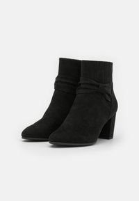 Bottines en daim noir avec un bout effilé, un détail noué décoratif à la cheville et un talon épais. Texture lisse.